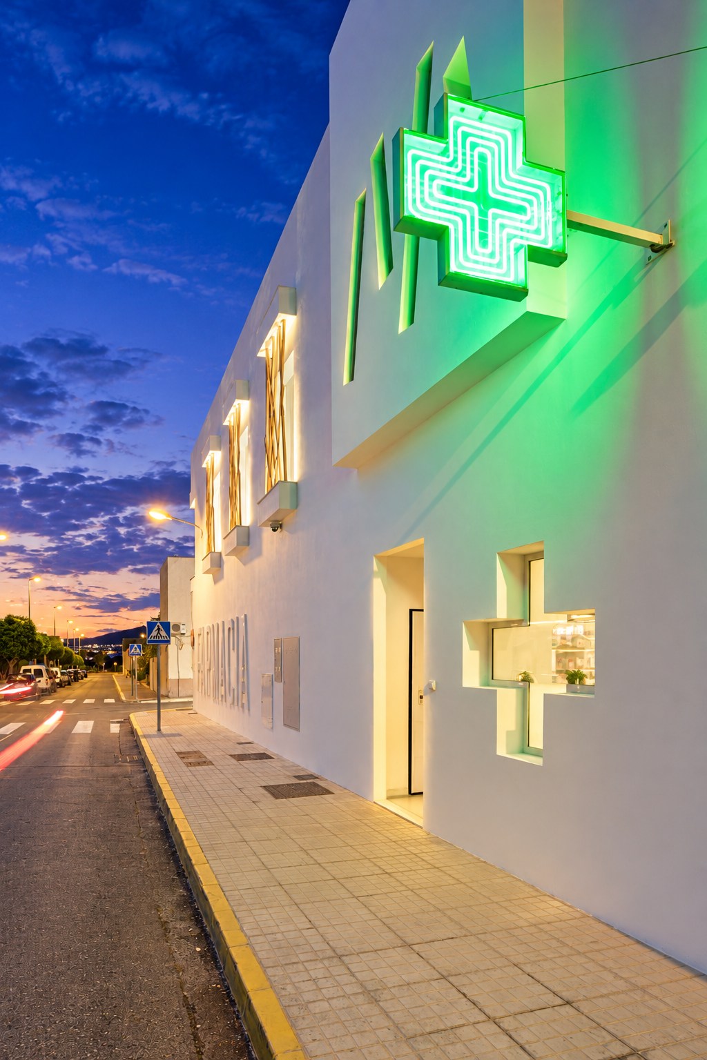 Vista lateral nocturna de Farmacia Caparrós y Reina con cruz luminosa y fachada blanca
